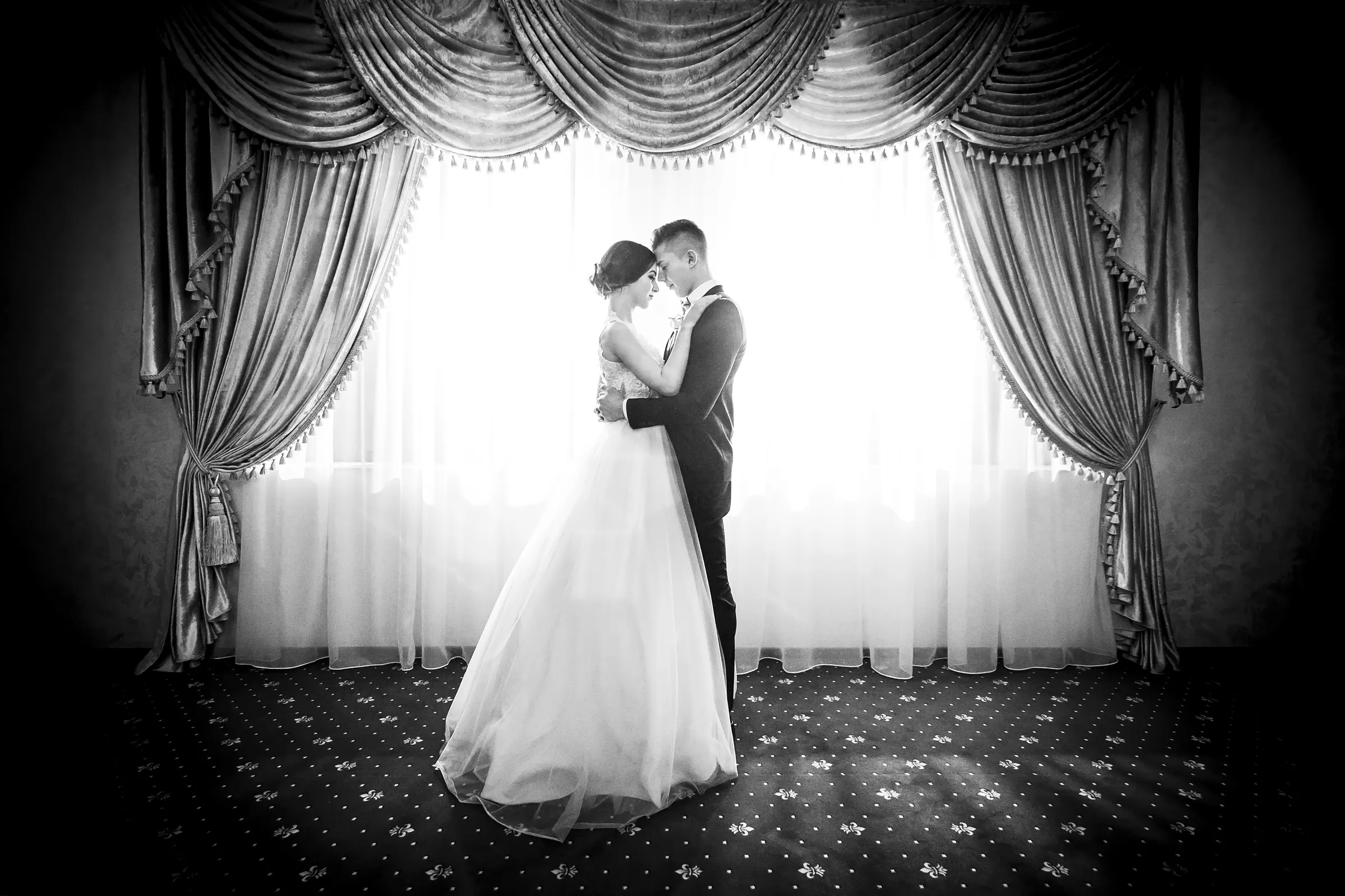 Bride and groom in a quiet embrace by a draped window, black and white